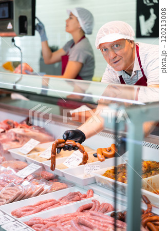 Male seller leans over to refrigerator window and takes out some raw barbecue sausages for buyer 120213008
