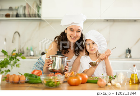 Mother and girl in white cooking hats are standing in kitchen near table with dishes. Mother and girl in white cooking hats are standing in kitchen near table with dishes. 120213844