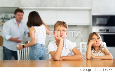 Two children sitting at the kitchen table sadly while their parents quarrelling Two children sitting at the kitchen table sadly while their parents quarrelling 120213977
