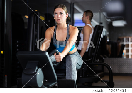 Sportive young girl training her arms using cable exercise machine in gym 120214314