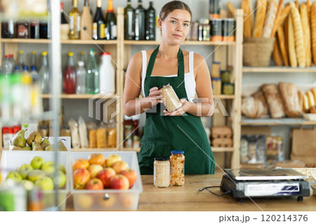 Portrait of friendly girl seller behind a store counter offering to buy canned beans and legumes Portrait of friendly girl seller behind a store counter offering to buy canned beans and legumes 120214376