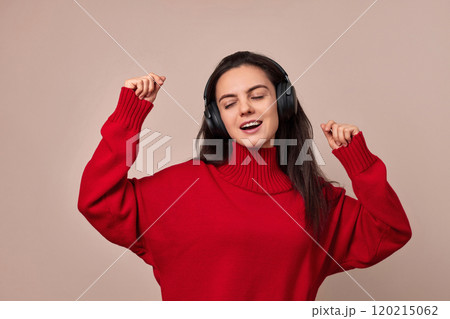 Happy woman wearing red sweater and headphones enjoying listening music and dancing on pink background. 120215062
