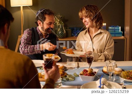 Jewish husband putting delicious food on wifes plate while they and their friends celebrating holiday together Jewish husband putting delicious food on wifes plate while they and their friends celebrating holiday together 120215259