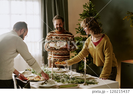 Caucasian family serving decorated table, they putting delicious dishes on white plates and appetizers during preparation for Christmas party 120215437