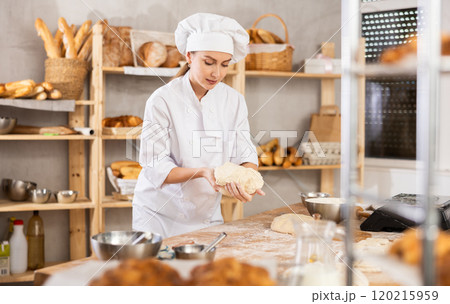 Skilled smiling young girl professional baker in white uniform standing at work table and kneading dough during working day in bakery 120215959