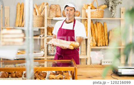 Middle-aged salesman holding paper bag with baguettes in bakery Middle-aged salesman holding paper bag with baguettes in bakery 120216099