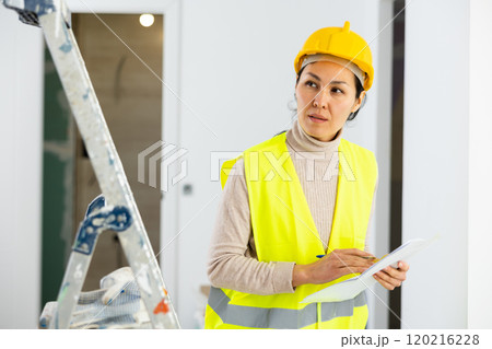 Woman foreman in a protective helmet and yellow vest checks the completed construction work on drawing Woman foreman in a protective helmet and yellow vest checks the completed construction work on drawing 120216228