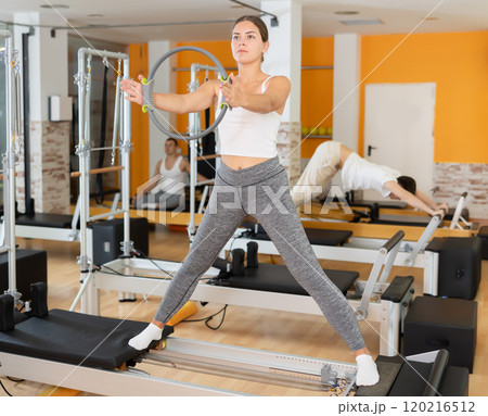 Girl stands on reformer and holds ring, hoop in hands, performs exercises to balance Girl stands on reformer and holds ring, hoop in hands, performs exercises to balance 120216512