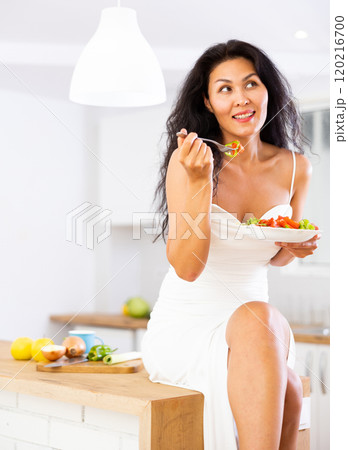 Woman sitting on kitchen table and eating salad Woman sitting on kitchen table and eating salad 120216700