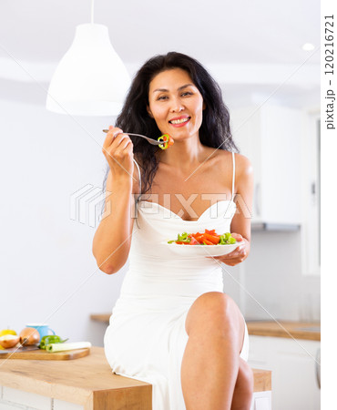 Woman sitting on kitchen table and eating salad Woman sitting on kitchen table and eating salad 120216721