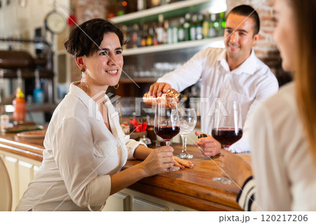 Two girlfriends chat with each other and drink red wine while sitting at bar in restaurant 120217206