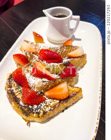 A delicious serving of French toast topped with fresh strawberries and a dusting of powdered sugar, served on a rectangular plate with a small pitcher of syrup on the side. The crispy, golden-brown 120217263