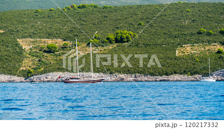 A peaceful sailing boat anchored near a lush green coastline with hills and blue waters. 120217332