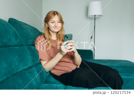 Beautiful blonde woman having fun online on her smartphone at home or hotel. Phone Communication and comfort concept. Smiling 30s woman daydreaming and holding mobile phone with copy space. 120217534