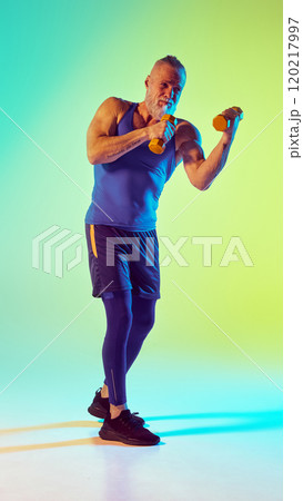 Kickboxing. Full-length of elderly sportive man training with dumbbell against gradient green yellow background in neon light Kickboxing. Full-length of elderly sportive man training with dumbbell against gradient green yellow background in neon light 120217997