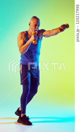 Kickboxing. Full-length of elderly sportive man training with dumbbell against gradient green yellow background in neon light Kickboxing. Full-length of elderly sportive man training with dumbbell against gradient green yellow background in neon light 120217998