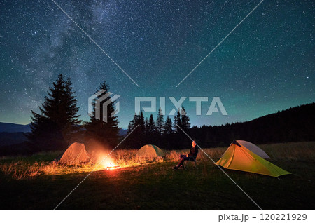 Person hiker having a rest by campfire, surrounded by glowing tents under magnificent starry sky. Milky Way stretches above, creating serene and magical atmosphere in peaceful mountain wilderness. Person hiker having a rest by campfire, surrounded by glowing tents under magnificent starry sky. Milky Way stretches above, creating serene and magical atmosphere in peaceful mountain wilderness. 120221929