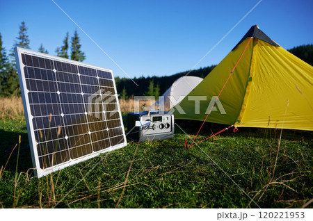 Tourist tent with solar panel and portable power station nearby in grassy field. Scene surrounded by rolling hills, tall pine trees under clear blue sky, showcasing eco-friendly camping setup. 120221953