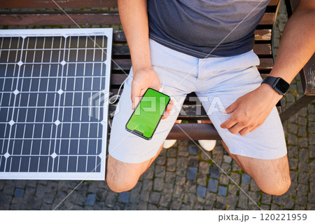 Young man using portable solar panel to charge his phone with green screen. Cropped photo of male sitting on bench and charging his mobile device by solar battery. Saving energy concept. 120221959