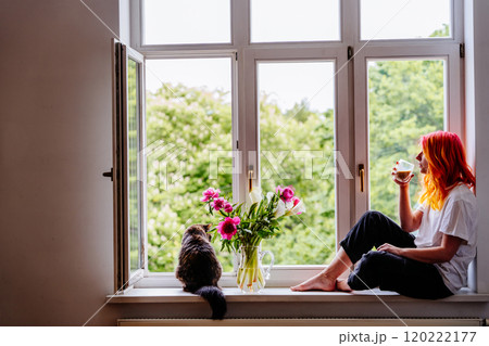 Young woman with bright hair sitting by large window, sipping coffee while enjoying tranquil view. Cat sitting nearby with vase of flowers, peaceful moments. 120222177