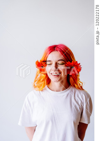 Portrait of smiling young Caucasian woman with vibrant pink and orange hair in white t-shirt, bright orange flowers behind her ears. Happiness, creativity, youthful energy, against clean background. Portrait of smiling young Caucasian woman with vibrant pink and orange hair in white t-shirt, bright orange flowers behind her ears. Happiness, creativity, youthful energy, against clean background. 120222178