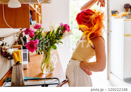 Vibrant young woman with colorful hair dances joyfully in bright kitchen with flowers on counter. Creativity and freedom, domestic art, joy, inspiring concept. 120222186