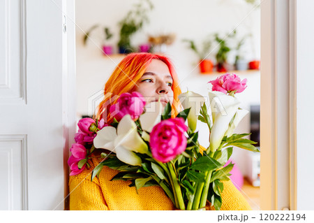 Portrait of young Caucasian woman with striking orange and yellow hair holding vivid bouquet of pink and white flowers at home. Soft focus of background shows blurred colorful potted plants. Portrait of young Caucasian woman with striking orange and yellow hair holding vivid bouquet of pink and white flowers at home. Soft focus of background shows blurred colorful potted plants. 120222194
