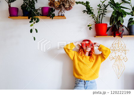 Young Caucasian woman with vibrant hair, bright yellow sweater, against white wall with plants on shelves, covering eyes with orange flowers. Creative energy, artistic vibe concept. Young Caucasian woman with vibrant hair, bright yellow sweater, against white wall with plants on shelves, covering eyes with orange flowers. Creative energy, artistic vibe concept. 120222199