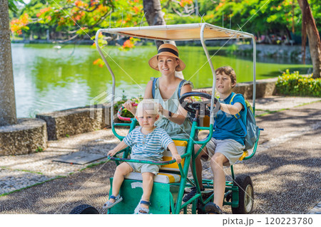 Mother tourist with her toddler and teenage sons riding an authentic Mexican pedal car in a park in Mexico. Family adventure and travel with kids concept 120223780