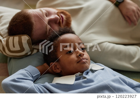Portrait shot of thoughtful little Black boy resting with adoptive father lying down on floor in living room while enjoying time together on weekend, copy space 120223927