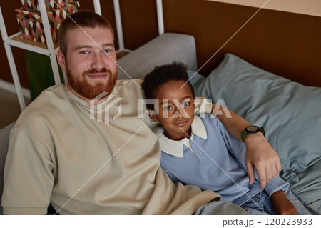 Portrait of smiling little Black boy looking at camera with happy adoptive dad while resting together sitting on couch in kids bedroom, copy space 120223933