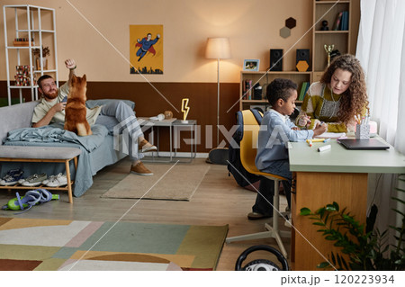 Wide angle shot of little Black boy sitting at desk with mom drawing pictures together in kids room while dad playing with dog in background, casual domestic life scene, copy space Wide angle shot of little Black boy sitting at desk with mom drawing pictures together in kids room while dad playing with dog in background, casual domestic life scene, copy space 120223934