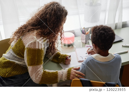 Candid shot of little Black boy and supportive mother engaged in hand drawing with colorful felt tip pens, while sitting together at desk in kids room 120223935