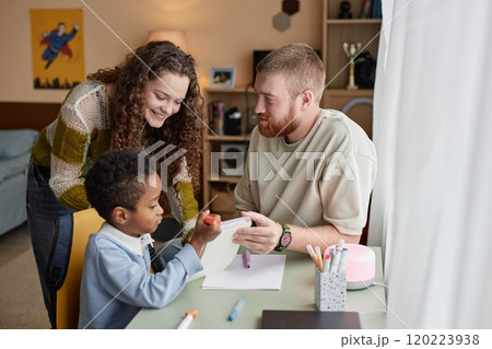 Medium shot of happy adoptive mother and father engaged in hand drawing with little Black boy sitting at desk in kids room 120223938