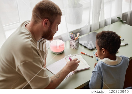 Candid shot of little Black boy and supportive father engaged in hand drawing with colorful felt tip pens, while sitting together at desk in kids room 120223940