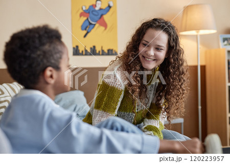 Medium shot of cheerful curly mother looking with adoring smile at little adopted kid while sitting on sofa in boys room 120223957