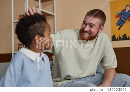 Medium shot of cheerful dad looking with adoring smile at little adopted kid praising son while sitting on sofa in boys room 120223961
