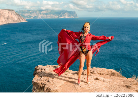 A woman in a red dress is standing on a rocky cliff overlooking the ocean. She is wearing a lei and she is dancing or celebrating. The scene is serene and peaceful, with the ocean as a backdrop. 120225080