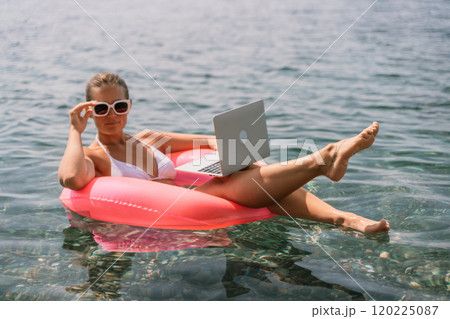 A woman is floating in a pink inflatable raft with a laptop in her hand. She is smiling and she is enjoying her time in the water. 120225087