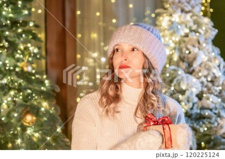 Woman Christmas Gift Lights - A woman wearing a white sweater and a blue beanie stands in front of a Christmas tree with lights. She is holding a gift wrapped in red paper with a bow. 120225124