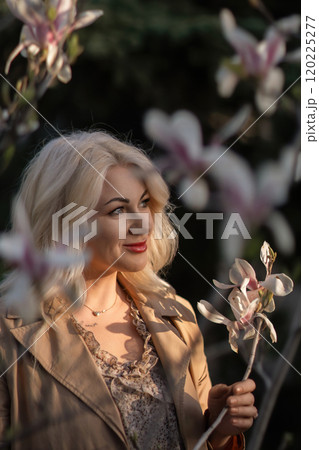 A woman is holding a magnolia flower in her hand and standing in front of a tree. Concept of serenity and beauty, as the woman is surrounded by nature and the flower adds a touch of color. 120225277