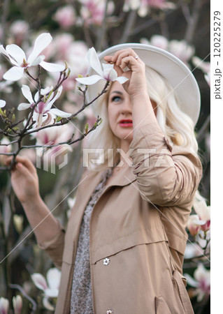 Magnolia flowers woman. A blonde woman wearing a white hat stands in front of a tree with pink flowers. She has a smile on her face and she is enjoying the beautiful scenery. Magnolia flowers woman. A blonde woman wearing a white hat stands in front of a tree with pink flowers. She has a smile on her face and she is enjoying the beautiful scenery. 120225279