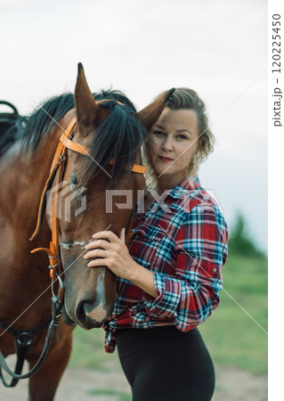 Happy blonde with horse in forest. Woman and a horse walking through the field during the day. Dressed in a plaid shirt and black leggings. Happy blonde with horse in forest. Woman and a horse walking through the field during the day. Dressed in a plaid shirt and black leggings. 120225450