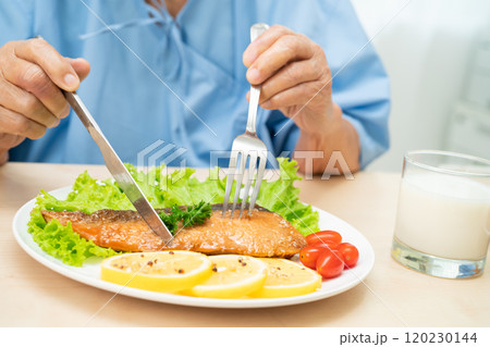 Asian senior woman patient eating Salmon steak breakfast with vegetable healthy food while sitting and hungry on bed in hospital. 120230144