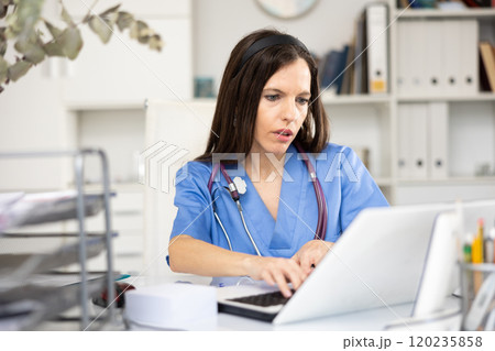 Woman doctor sitting at table in her office and using laptop 120235858