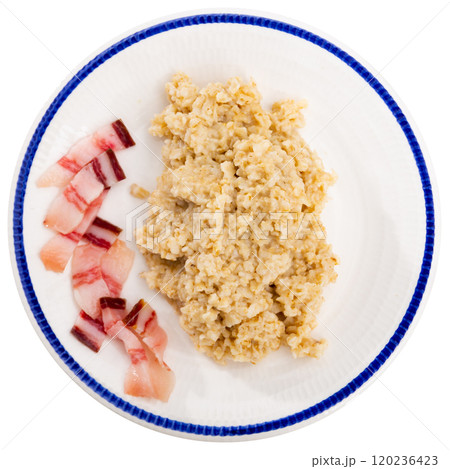 There is portion of boiled oatmeal porridge with pieces of salted bacon on plate. 120236423