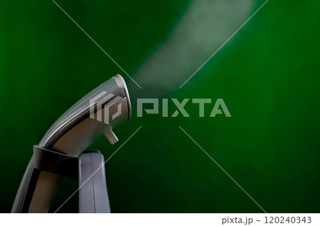 A gray and white steam generator stands close-up on a green background, a large stream of steam streams from it on a green background with space for text. Ironing concept, steam treatment and gentle A gray and white steam generator stands close-up on a green background, a large stream of steam streams from it on a green background with space for text. Ironing concept, steam treatment and gentle 120240343