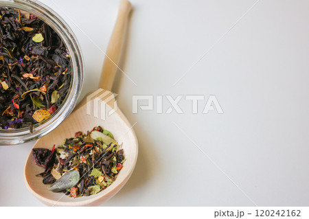 Large-grain tea with the addition of pieces of fruit and berries lies on a wooden spoon, next to it is a tilted glass jar with tea on a white background with space for text, Concept of tea drinking 120242162