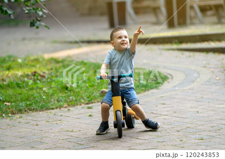 A cute smiling toddler boy of three or four years old rides a bicycle or balance bike in a city park on a sunny summer day. Toddlerhood and childhood concept. Selective focus 120243853