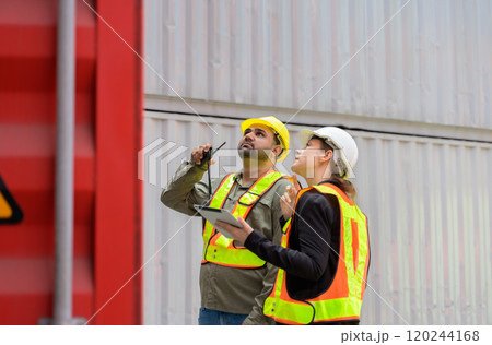 Foreman man with yellow helmet holding walkie talkie and working with female manager on digital tablet while standing in front of red openning container box at cargo shipping yard. 120244168